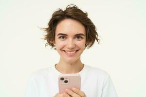 tecnología concepto. joven mujer utilizando su móvil teléfono, participación teléfono inteligente y sonriente, utilizando aplicación, hojeada social medios de comunicación, haciendo en línea compras, aislado terminado blanco antecedentes foto