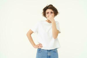 Portrait of woman disgusted by bad smell, shuts her nose, stands over white studio background photo