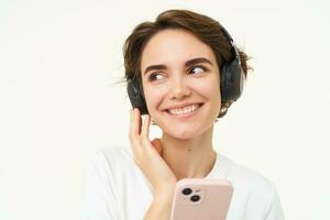 Portrait of candid young woman, listens music in wireless headphones, holding smartphone, watching videos on mobile phone, standing over white background photo