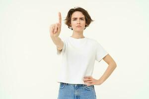 Portrait of serious young woman, shows one finger, stop gesture, disapprove something with frowning face, stands over white background photo
