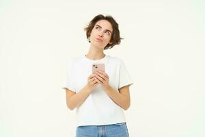 Portrait of thinking girl standing with smartphone, looking up thoughtful, making choice while buying smth online, placing an order, standing over white background photo