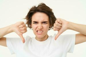 Close up portrait, face of young woman that is showing thumbs down, dislike, do not recommend gesture, express negative feedback, isolated over white background photo