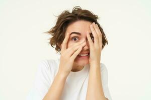 Image of funny woman peeking through fingers, looking at something with curious face expression, waiting for surprise and smiling excited, isolated over white background photo