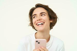 retrato de joven mujer en pie con rosado móvil teléfono, utilizando teléfono inteligente aplicación, haciendo en línea compras, pago para alguna cosa utilizando solicitud, en pie terminado blanco antecedentes foto