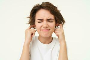 Young woman shuts her ears from loud noise, hears siren, distressed by shouting neighbours, standing over white background photo