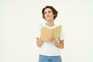 Portrait of thoughtful and creative woman writing down her thoughts in diary, doing homework, looking aside and thinking, posing over white background photo