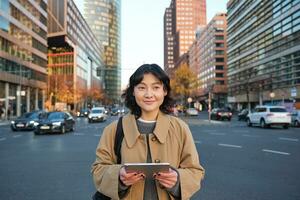 Portrait of smiling korean girl stands on busy street of city centre, holds digital tablet, casual relaxed face expression, walking around town photo