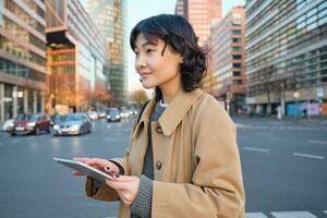 Woman walking on street using digital tablet, holding gadget in hands and looking away, standing in city centre photo