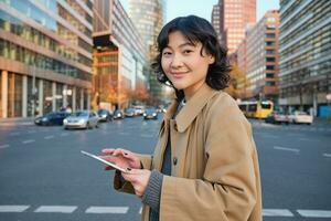 Woman walking on street using digital tablet, holding gadget in hands and looking away, standing in city centre photo