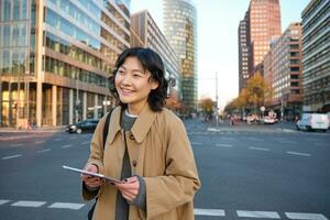 Portrait of smiling korean girl stands on busy street of city centre, holds digital tablet, casual relaxed face expression, walking around town photo