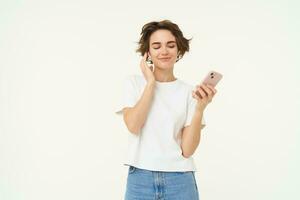 Portrait of happy brunette woman, holds smartphone, listens music in wireless headphones, stands over white studio background photo
