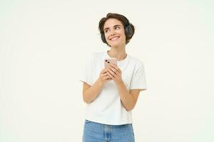 Image of happy young girl listens to music in headphones, standing with smartphone, using mobile phone streaming app, white background photo