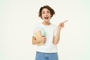 Portrait of brunette woman laughing, student with notebooks pointing at upper right corner, showing banner or advertisement, standing over white studio background photo