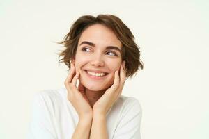 Close up of hopeful young woman, girl anticipating something, touching her face, looking forward to something, waiting with excitement, standing over white background photo