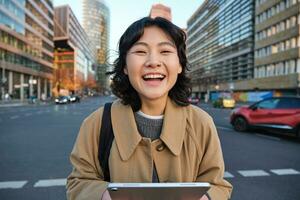 Portrait of asian girl student, stands in city centre with cars on busy street, holds digital tablet and smiles at camera photo