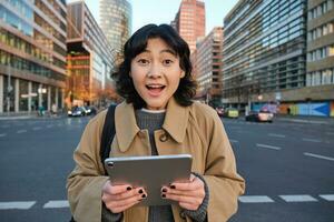 Portrait of korean girl looks surprised, found out something amazing, holding digital tablet with joyful face, stands on street of city centre photo