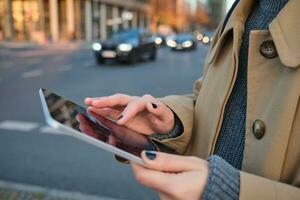 Close up photo of female hands holding digital tablet, girl looks at screen, checks application, stands on busy street filled with cars in afternoon