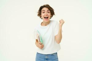 Enthusiastic brunette woman makes fist pump, holds documents and notes, looks thrilled and happy, winning, triumphing, posing over white background photo