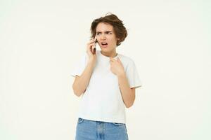 Portrait of shocked, angry woman talks on mobile phone with disappointed, furious face expression, standing over white background photo