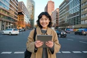 Portrait of asian girl student, stands in city centre with cars on busy street, holds digital tablet and smiles at camera photo