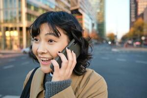 retrato de sorprendido coreano chica, negociaciones en móvil teléfono, oye increíble Noticias terminado teléfono conversación, soportes en calles de ciudad con emocionado cara expresión foto