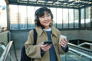 Portrait of smiling korean girl drinks coffee to go, goes up an escalator, holds smartphone, visits new city, arrives at train station, listens music in headphones photo