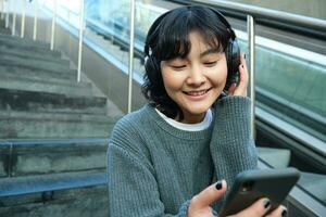 retrato de sonriente coreano niña en auriculares, usos teléfono inteligente y se sienta en escalera en centro comercial, relojes vídeo en móvil teléfono foto