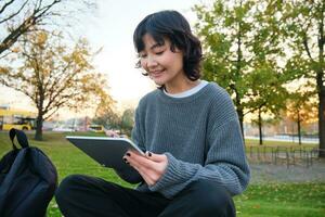 Portrait of young smiling korean girl, graphic designer, artist drawing on digital tablet with a pen tool, sitting in park on fresh air and scatching, taking notes photo