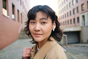 Stylish modern asian girl, student in wireless headphones, walks around city with backpack, smiling with happy face photo