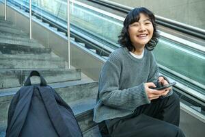 Stylish young girl student, sits on stairs with smartphone and backpack, laughs and smiles, texts message, uses social media photo