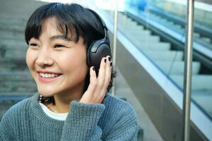 Portrait of smiling korean girl in headphones, uses smartphone and sits on stairs in mall, watches video on mobile phone photo