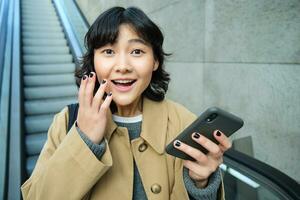 alegre y positivo coreano chica, celebra, mira sorprendido, va abajo escalera mecánica con teléfono inteligente y mira asombrado por algo foto