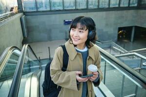 Portrait of smiling korean girl drinks coffee to go, goes up an escalator, holds smartphone, visits new city, arrives at train station, listens music in headphones photo