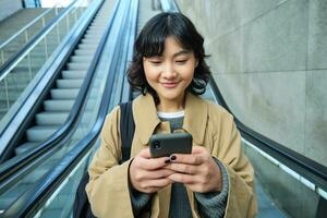 Portrait of beautiful asian girl, student goes down escalator in city, looks at mobile phone, uses telephone, map application, commutes somewhere in town photo