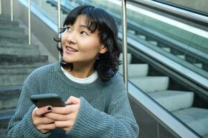 Stylish young girl student, sits on stairs with smartphone and backpack, laughs and smiles, texts message, uses social media photo