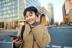 Portrait of young asian girl, student walks in city, listens music in headphones and uses mobile phone on streets photo