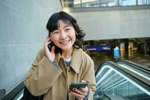 retrato de linda sonriente coreano chica, va arriba escalera mecánica, escucha música en inalámbrico auriculares y usos móvil teléfono, sostiene teléfono inteligente foto