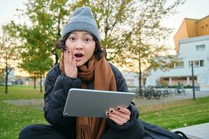 Portrait of asian girl with shocked, surprised face, holds tablet, read or watched big news, looks amazed, sits in park on bench, wears warm clothes photo