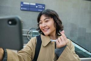 Portrait of stylish korean girl, student takes selfie on street, holds smartphone and poses on its camera, makes video on tourist trip, smiles with joy photo