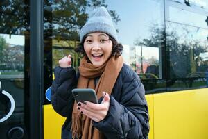 Enthusiastic girl rejoice, reads message on mobile phone and celebrates, stands near her bus on public transport stop and looks excited, posing in warm winter clothes photo