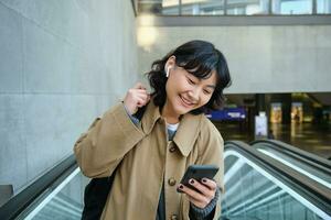 Portrait of cute korean girl in trench coat, going up escalator, holds mobile phone and smiles pleased, commutes, watches video or listens music with wireless headphones photo