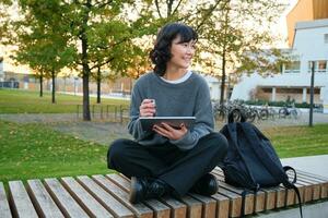 Young asian girl, student sits on bench, graphic designer with pencil and digital tablet draws scatches, does her homework outdoors photo