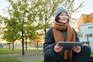 Portrait of asian girl in warm clothes, sits on bench with digital tablet and graphic pen, smiling happily, draws outdoors in chilly weather photo