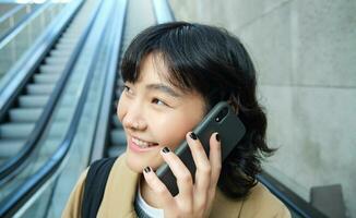 disparo a la cabeza de sonriente coreano mujer con teléfono inteligente, hace un teléfono llamar, va abajo escalera mecánica en ciudad, viaja a Universidad foto