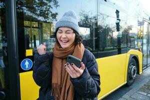 moderno personas y estilo de vida. contento asiático niña carcajadas desde alegría, celebra, soportes cerca autobús público transporte y mira asombrado foto