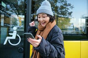 moderno personas y estilo de vida. contento asiático niña carcajadas desde alegría, celebra, soportes cerca autobús público transporte y mira asombrado foto