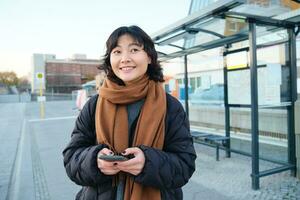 contento sonriente coreano chica, utilizando móvil teléfono, en pie en autobús detener con teléfono inteligente, mirando a salida calendario en solicitud, posando en invierno ropa foto