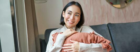 Women and wellbeing concept. Young woman in warm clothes, sits at home, hugs pillow on sofa and smiles pleased, thinks of smth photo