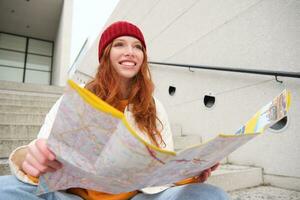 joven sonriente pelirrojo chica, turista se sienta en escalera al aire libre con ciudad papel mapa, mirando para dirección, viajero mochilero explora ciudad y mira para Turismo foto