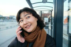 cerca arriba de linda coreano mujer, haciendo un teléfono llamar, hablando y sonriente en teléfono, en pie en invierno chaqueta en autobús detener, esperando para su transporte a llegar foto